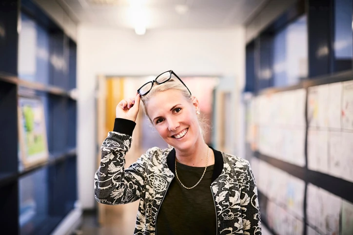 Smiling person with short blond hair holds glasses on head, standing in a hallway with artwork displayed on the wall.