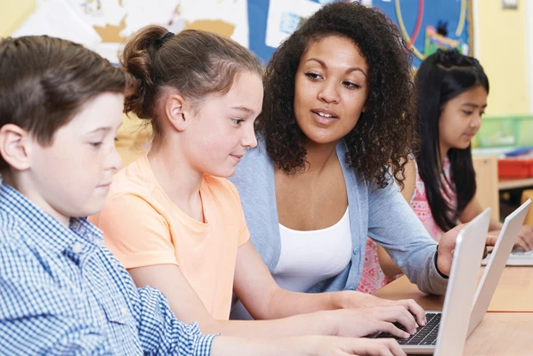 Teacher helping students with laptops in a classroom; diverse group focused on their devices.