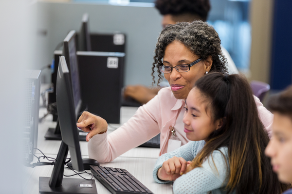 An educator, sitting in front of a computer, assists a student with classwork