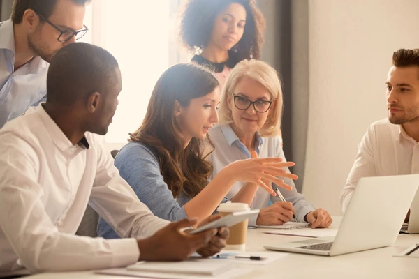 Diverse group of colleagues in a meeting room, discussing around a table with a laptop, notes, and coffee cup.