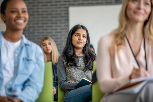 A group of diverse students attentively listening in a classroom setting, with focus on a woman in the center holding a notebook.