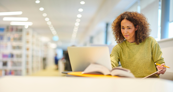 A woman sits in a library in front of her laptop,...