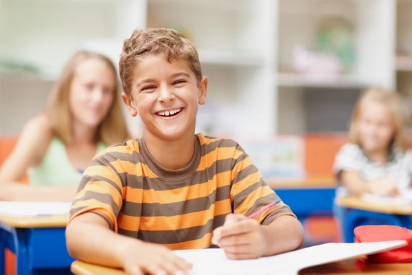 Boy in striped shirt smiling, seated at desk in a classroom, with blurred students in the background.