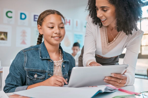 A teacher showing a tablet to a student in a classroom. The student smiles while looking at the screen.