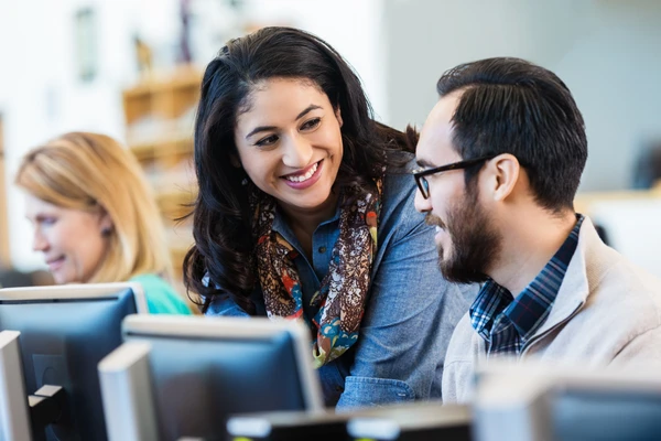 Two people smiling and talking while working on computers in an office setting. A third person is focused on their screen.