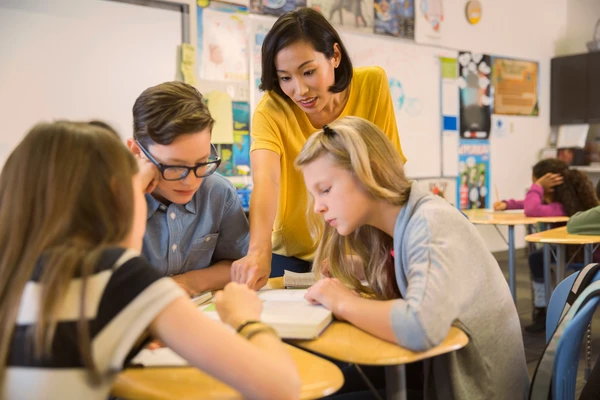 A teacher helps three students with a book at their desk in a classroom setting.