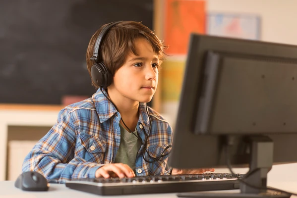 Young boy wearing headphones, focused on a computer screen, seated at a desk with a keyboard and mouse.