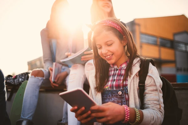 A girl in overalls smiles while looking at a tablet outdoors, with two other people in the background during a sunny day.