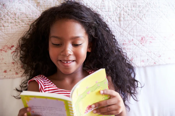 Girl with long hair reading a yellow book while lying on a bed with a floral quilt.