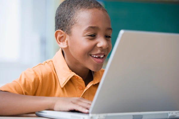 A smiling child in an orange shirt uses a laptop indoors, appearing engaged and happy.