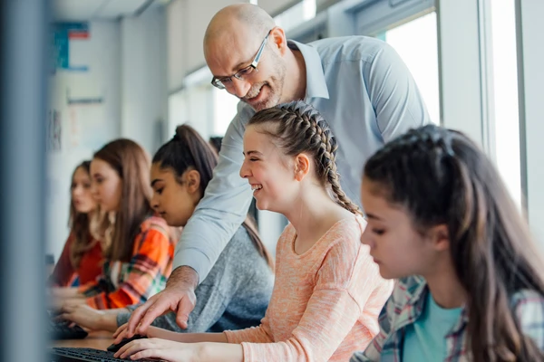 A teacher assists a smiling student on a computer in a classroom, surrounded by other focused students.