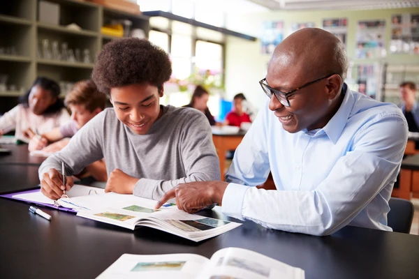 A teacher and student smiling while discussing a textbook in a classroom. Other students are visible in the background.