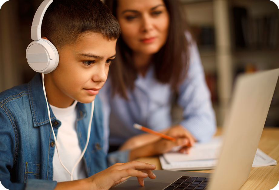 A student wearing headphones looks at a laptop while completing schoolwork as a teacher sits behind him and monitors his progress,