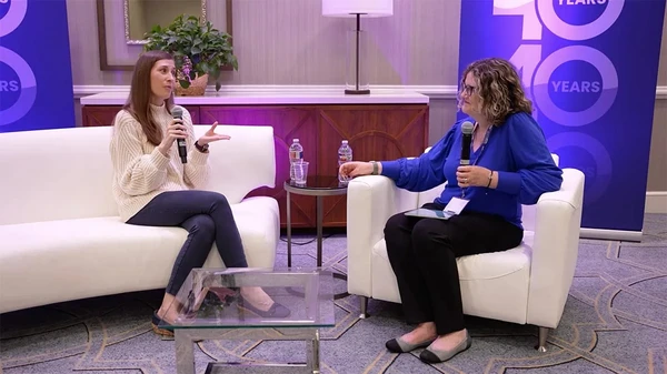 Two women sit on white couches, talking with microphones at an indoor event with 10 Years banners in the background.