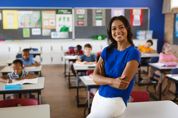 An educator with hands folded leans on a desk in front of a classroom while students are busy working